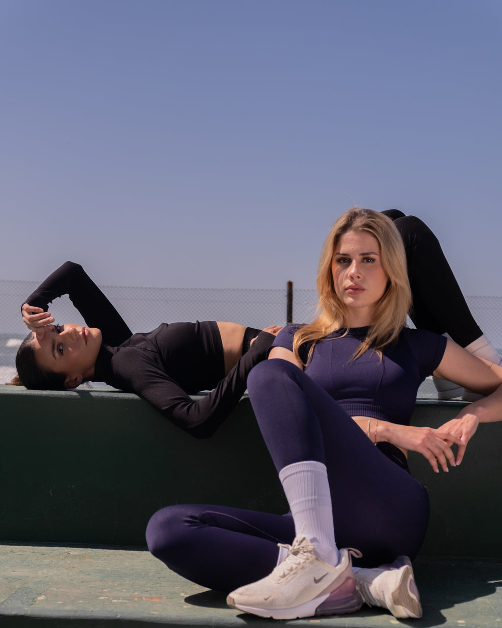Two women in athletic wear sitting on a ledge with a clear blue sky background