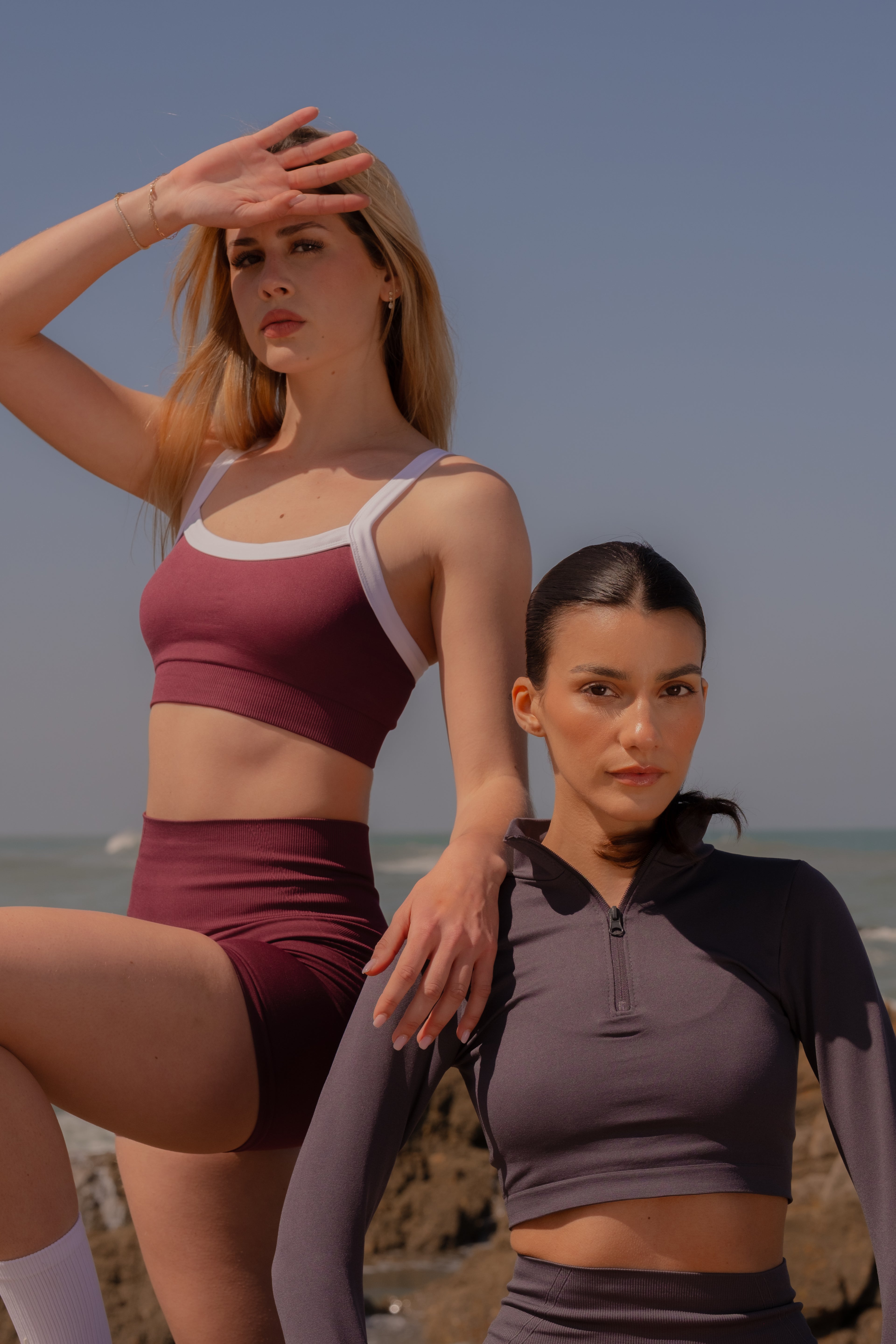 Two women in athletic wear standing on a beach with ocean and sky in the background.